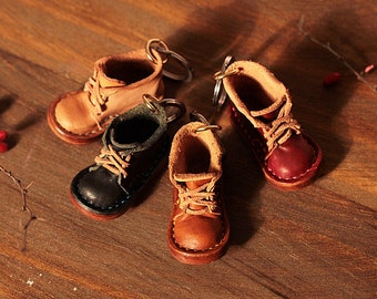 Puede incluir: Cuatro llaveros de bota de cuero en miniatura en varios colores: marrón claro, negro, marrón y burdeos. Cada bota tiene cordones y un llavero de metal. Las botas están dispuestas sobre una superficie de madera.
