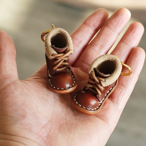 May include: A pair of brown leather miniature boots with tan fur lining and white stitching. The boots are attached to a gold key ring.