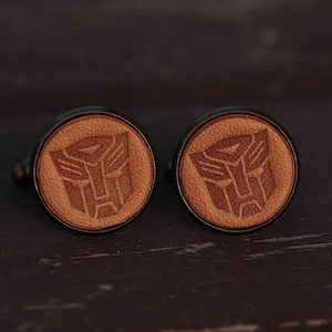 May include: A pair of brown leather cufflinks with a black metal frame. The cufflinks feature a laser-engraved image of the Transformers logo.
