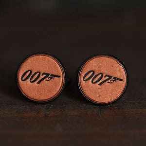 Op de afbeelding: Een paar ronde manchetknopen met een bruin, leerachtig oppervlak. Elke manchetknop toont het nummer "007" met een zwart pistoolsilhouet. De manchetknopen hebben een zwarte rand.