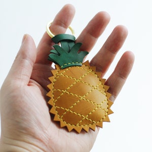 May include: A yellow leather pineapple keyring with green leather leaves and a gold metal ring. The pineapple is stitched with yellow thread.