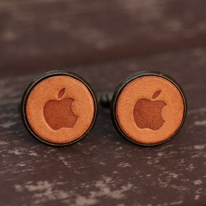 May include: A pair of brown leather cufflinks with a silver-toned metal backing. Each cufflink features a raised, embossed apple logo.