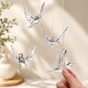 May include: Four clear, faceted crystal hummingbird ornaments suspended by thin silver strings. Each bird is depicted in a different flight pose with wings spread wide, showcasing intricate geometric cuts that reflect light and create a sparkling effect.
