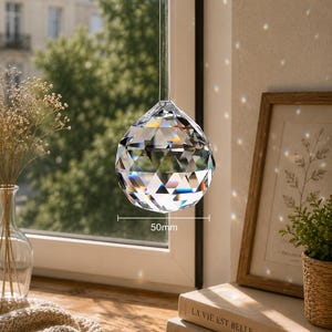 May include: A clear, multi-faceted crystal sphere hanging by a thin cord. The crystal features a geometric cut that refracts light, creating small rainbow spots. A label indicates a diameter of 50mm. The object is suspended in front of a window.