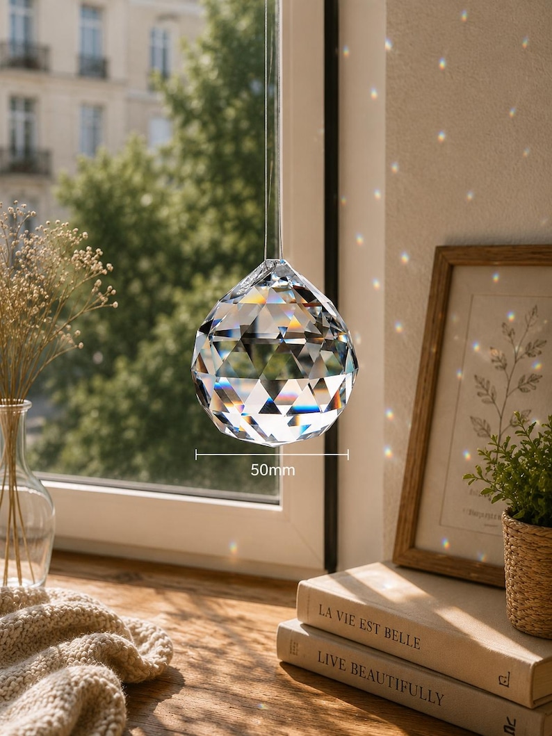 May include: A clear, multi-faceted crystal sphere hanging by a thin cord. The crystal features a geometric cut that refracts light, creating small rainbow spots. A label indicates a diameter of 50mm. The object is suspended in front of a window.
