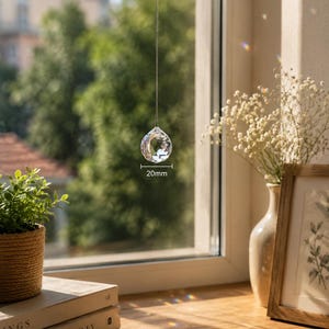May include: A clear, multifaceted crystal sphere hanging from a thin string in front of a window. The crystal has a diameter of 20mm as indicated by the text and measurement lines visible on the object, creating light refractions on the surface.