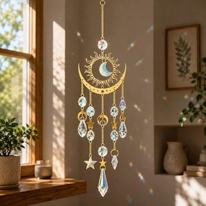 May include: A gold-toned metal sun and moon hanging ornament featuring a central crescent moon and sunburst design. The piece is adorned with multiple clear faceted crystal prisms, small gold stars, and additional crescent moon charms on chains.
