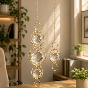 May include: Two hanging ornaments featuring faceted clear crystal spheres suspended within gold-toned twisted metal rings. The longer ornament contains three crystal spheres, while the shorter one contains one, both attached to thin gold-toned chains.
