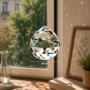 May include: A clear, multi-faceted crystal sphere hanging in front of a window. The crystal features a geometric cut that refracts light, creating small rainbow spots on the wall. A measurement line indicates a diameter of 60mm.
