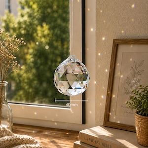 May include: A clear, multifaceted crystal suncatcher sphere hanging in front of a window. The sphere features a geometric cut design and is labeled with a 40mm measurement indicator. It refracts sunlight, casting small, colorful light spots.