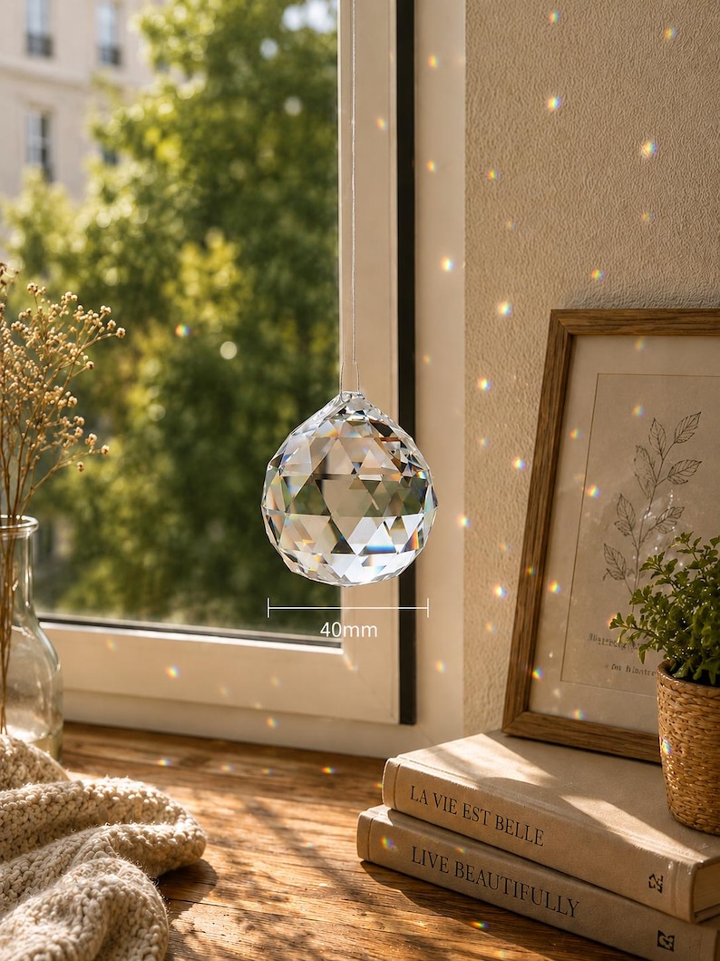 May include: A clear, multifaceted crystal suncatcher sphere hanging in front of a window. The sphere features a geometric cut design and is labeled with a 40mm measurement indicator. It refracts sunlight, casting small, colorful light spots.