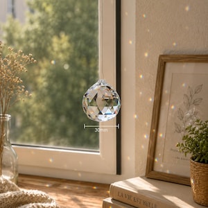 May include: A clear, multi-faceted crystal sphere hanging in front of a window, casting colorful light refractions. The crystal features a geometric cut and is labeled with a 30mm measurement indicator, showing its diameter clearly in the center.