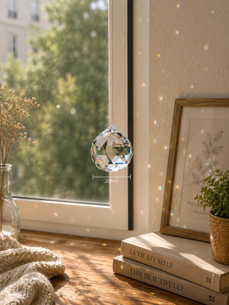 May include: A clear, multi-faceted crystal sphere hanging in front of a window, casting colorful light refractions. The crystal features a geometric cut and is labeled with a 30mm measurement indicator, showing its diameter clearly in the center.