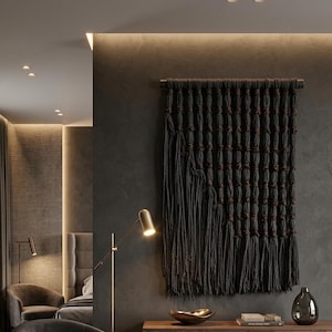 May include: A large rectangular wall hanging made of thick dark charcoal gray cord, featuring a grid of vertical columns with knotted sections and long loose fringe at the bottom, suspended from a horizontal wooden rod mounted on a dark wall.