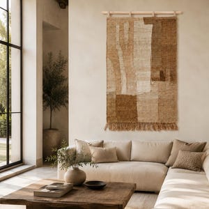 May include: A rectangular wall hanging tapestry featuring a textured, woven pattern in shades of beige, tan, and dark brown. The piece is suspended from a light-coloured wooden rod with fabric loops and features a fringed bottom edge.