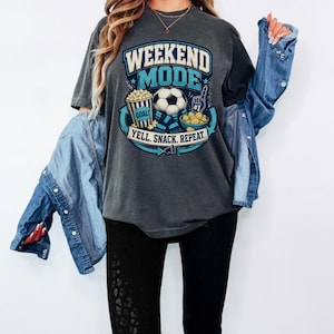 Op de afbeelding: Een antracietgrijs t-shirt met korte mouwen en een grafische print met de tekst WEEKEND MODE en YELL. SNACK. REPEAT. Het ontwerp bevat een voetbal, een popcornemmer met het label GOAL, een schaal met snacks en een nummer &eacute;&eacute;n schuimvinger.