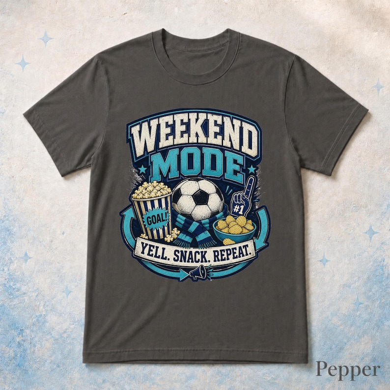 Op de afbeelding: Een antracietgrijs t-shirt met een afbeelding met de tekst WEEKEND MODE en YELL. SNACK. REPEAT. Het ontwerp bevat een voetbal, een popcornemmer met het label GOAL, een schaal met snacks en een nummer &eacute;&eacute;n schuimvinger in blauw en wit.