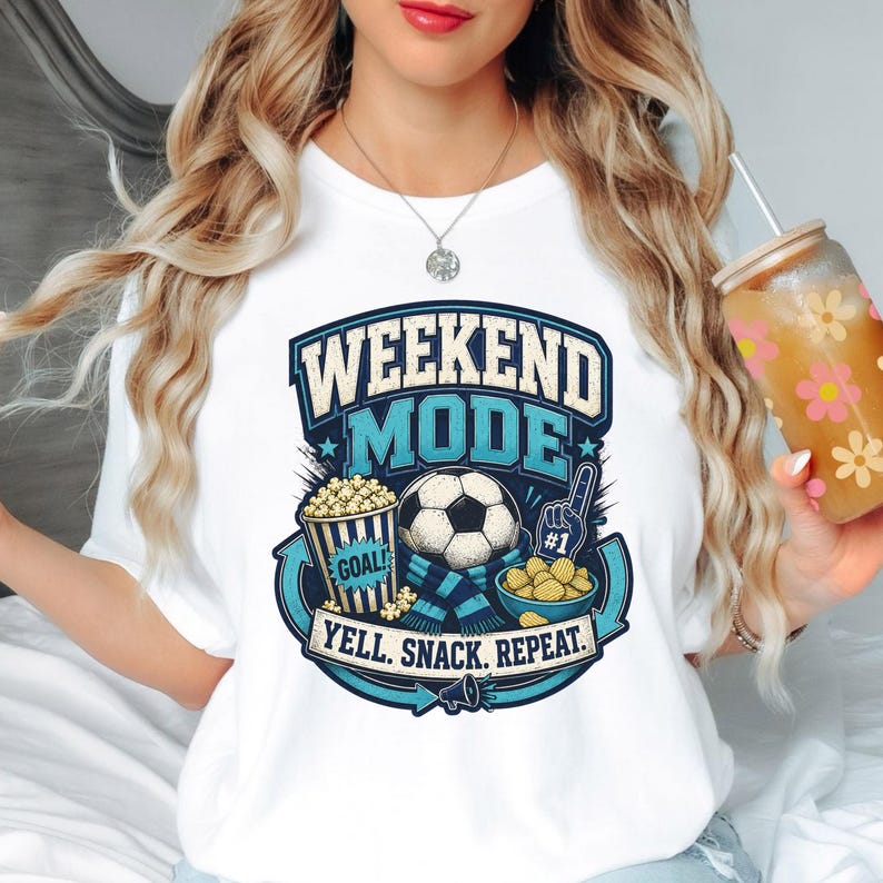 Op de afbeelding: Een wit t-shirt met een grafisch ontwerp met de tekst WEEKEND MODE en YELL. SNACK. REPEAT. De illustratie bevat een voetbal, een emmer popcorn met het label GOAL, een schaal met snacks en een blauwe en witte schuimvinger met nummer &eacute;&eacute;n.