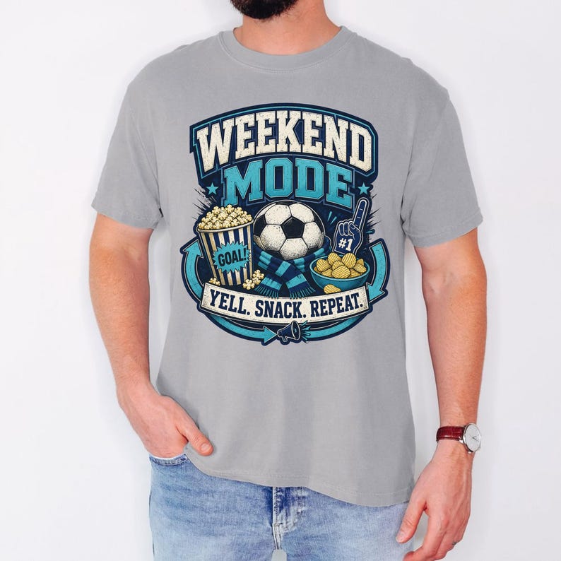 Op de afbeelding: Een lichtgrijs t-shirt met een afbeelding met de tekst WEEKEND MODE en YELL. SNACK. REPEAT. Het ontwerp bevat een voetbal, een emmer popcorn met het label GOAL, een schaal met snacks, een blauwe sjaal en een nummer &eacute;&eacute;n schuimvinger.