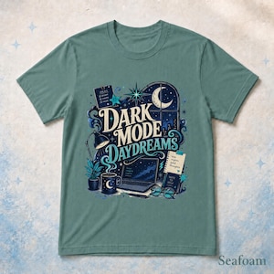 May include: A sage green t-shirt featuring a graphic illustration with the text "DARK MODE DAYDREAMS". The design includes a laptop, a desk lamp, a mug with a crescent moon, a notepad, glasses, and a window showing a night sky with a moon.
