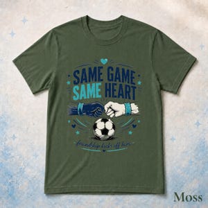 May include: An olive green short-sleeve t-shirt featuring a graphic of two fists bumping above a soccer ball. The text reads SAME GAME SAME HEART and friendship kicks off here. The design includes small blue stars and a heart icon.