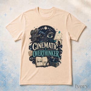 May include: A light beige t-shirt featuring a graphic print with the text "CINEMATIC OVERTHINKER". The design includes a vintage film camera, a large eye, a notebook, a stack of books, a coffee mug, and film strips in shades of blue and black.