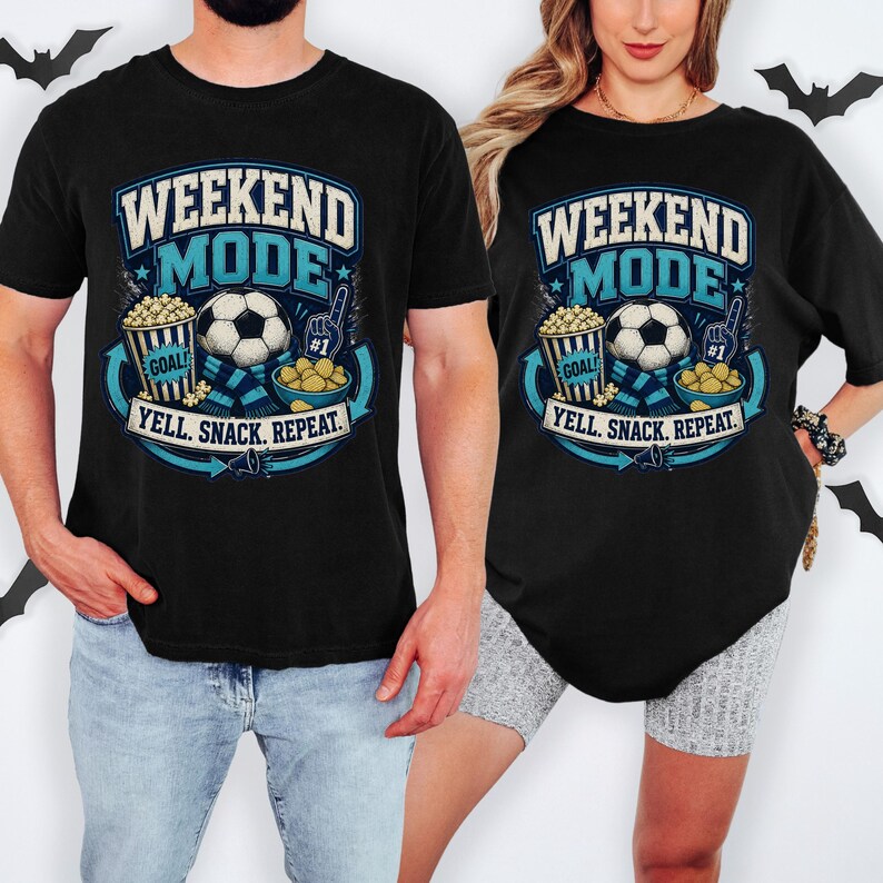 Op de afbeelding: Een zwart t-shirt met een afbeelding met de tekst WEEKEND MODE en YELL. SNACK. REPEAT. Het ontwerp bevat een voetbal, een popcornemmer met het label GOAL, een schaal met snacks en een schuimvinger, uitgevoerd in blauw, wit en geel.
