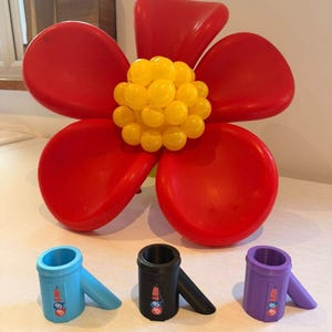 May include: A large flower sculpture made of red and yellow balloons, accompanied by three small cylindrical plastic connectors in light blue, black, and purple. Each connector features a small logo with the text UP UP & AWAY.