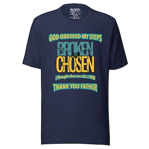 May include: A navy blue short-sleeve t-shirt featuring the text GOD ORDERED MY STEPS, BROKEN BUT CHOSEN, I thought that was ALL I HAD, and THANK YOU FATHER in yellow and teal lettering. The shirt has a crew neckline and a Rockwater brand label.