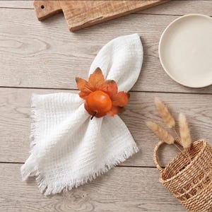 May include: A white waffle-textured napkin with frayed edges held by a decorative napkin ring featuring a bright orange pumpkin and several brown and orange autumn leaves. The napkin is folded and positioned on a light-colored wooden surface.