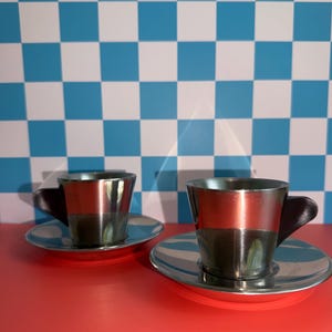 Puede incluir: Dos tazas de espresso metálicas con asas de color marrón oscuro, cada una sobre un platillo circular plateado a juego. Las tazas presentan un acabado pulido y reflectante con una forma cónica para servir pequeñas porciones de café.