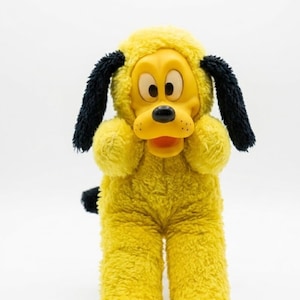 May include: A plush toy of a yellow dog with a molded plastic face, large white eyes, a black nose, and an open mouth. The body is covered in yellow fuzzy fabric, featuring long black floppy ears and a small black tail extending from the back.