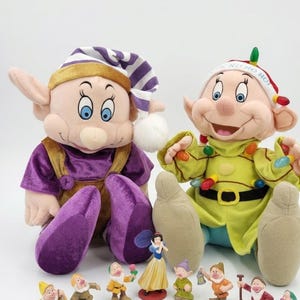 May include: Two plush dolls of Dopey from Snow White, one wearing a purple outfit and striped hat, the other in a green outfit with Christmas lights, accompanied by eight small figurines of Snow White and the seven dwarfs on a white surface.