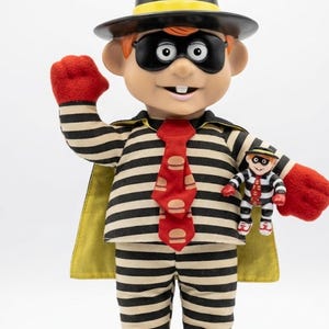 Op de afbeelding: Een pluche pop van een personage met een zwart-wit gestreept shirt en broek, een zwart masker, een zwarte hoed met een gele band, een rode das met hamburgerpictogrammen, rode handschoenen en witte schoenen, met een kleiner bijpassend figuurtje.