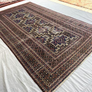May include: A rectangular area rug featuring a dark brown field with a central series of geometric diamond motifs in navy blue, cream, and rust red. The rug is framed by multiple intricate borders with repeating patterns in matching earth tones.