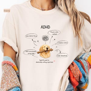 May include: An ivory t-shirt featuring a graphic of a golden retriever holding a stick in its mouth. The text reads ADHD, was told to focus, found a stick instead, mission: critical, 10/10 commitment, wrong task, task: abandoned, and hyperfocused on absolutely nothing important.