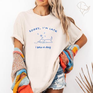 以下が含まれることがあります： SORRY, I'M LATE i saw a dogという青い文字と、背中に花を乗せて横たわる犬と骨の線画が描かれたベージュの半袖Tシャツ。カラフルなニットカーディガンを羽織っています。