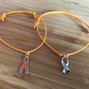 May include: Two orange string bracelets with ribbon charms. One charm is an orange ribbon, the other is a silver ribbon with the word "HOPE". The bracelets are on a wooden surface.
