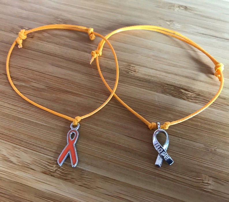 May include: Two orange string bracelets with ribbon charms. One charm is an orange ribbon, the other is a silver ribbon with the word "HOPE". The bracelets are on a wooden surface.