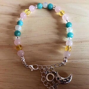 May include: A bracelet made with pink, yellow, and turquoise beads with a silver charm of a crescent moon and a female symbol.