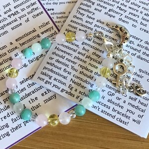 May include: A bracelet with pink, yellow, and green beads, a silver charm with a crescent moon and stars, and a silver charm with a female symbol. The bracelet is on a white card with text about emotional roller coasters and gemstones.