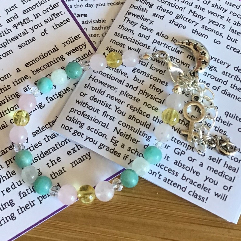 May include: A bracelet with pink, yellow, and green beads, a silver charm with a crescent moon and stars, and a silver charm with a female symbol. The bracelet is on a white card with text about emotional roller coasters and gemstones.