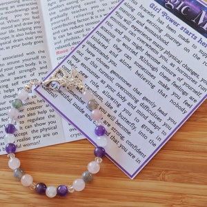 May include: A bracelet with pink, purple, and gray beads and a silver charm. The bracelet is accompanied by a card that reads "Girl Power starts here" and includes a message about overcoming challenges and finding strength.