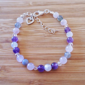 May include: A silver bracelet with a heart charm features a gradient of amethyst, moonstone, and rose quartz beads.