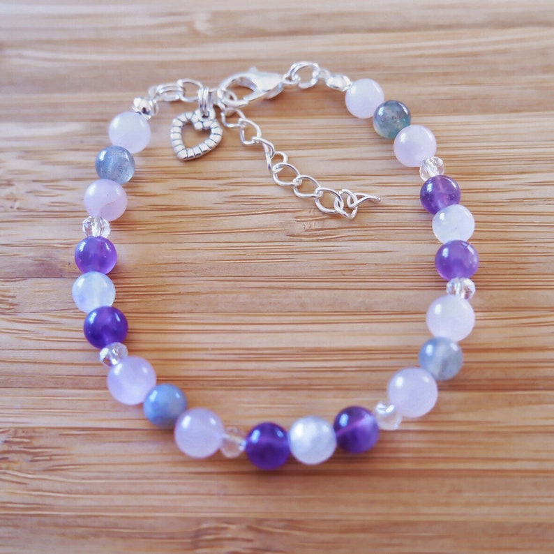 May include: A silver bracelet with a heart charm features a gradient of amethyst, moonstone, and rose quartz beads.