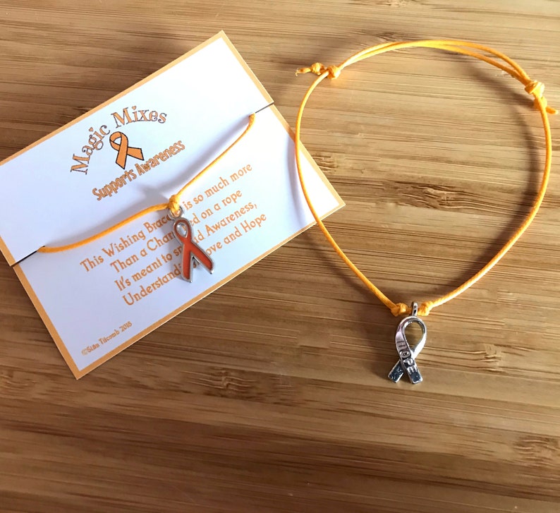 May include: An orange awareness ribbon charm bracelet with a matching card. The card reads "Magic Mixes Supports Awareness" and includes the text "This Wishing Bracelet is so much more than a charm on a rope..." The bracelet is on a wooden surface.