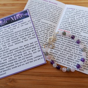 May include: A bracelet made with amethyst, rose quartz, and other gemstones. The bracelet is on a wooden surface with a card that reads "Girl Power Mixes" and describes the properties of each gemstone.