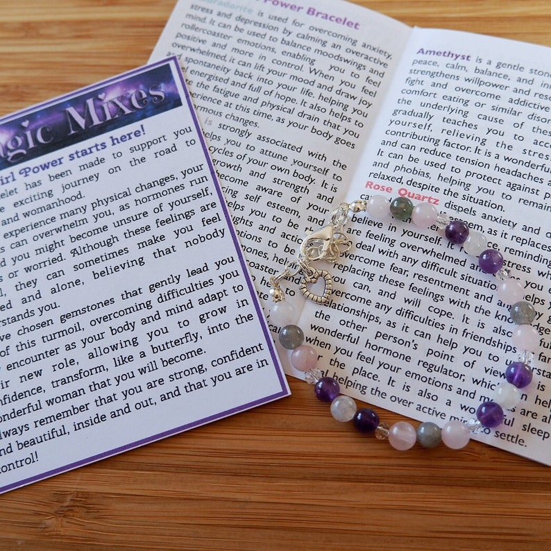 May include: A bracelet made with amethyst, rose quartz, and other gemstones. The bracelet is on a wooden surface with a card that reads "Girl Power Mixes" and describes the properties of each gemstone.