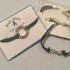 May include: Two teal suede bracelets with silver charms. One bracelet features a round charm with the word "SURVIVOR" engraved around it. The other bracelet features an infinity symbol charm. The bracelets are packaged on white cards with the text "Magic Mixes Supports Awareness".