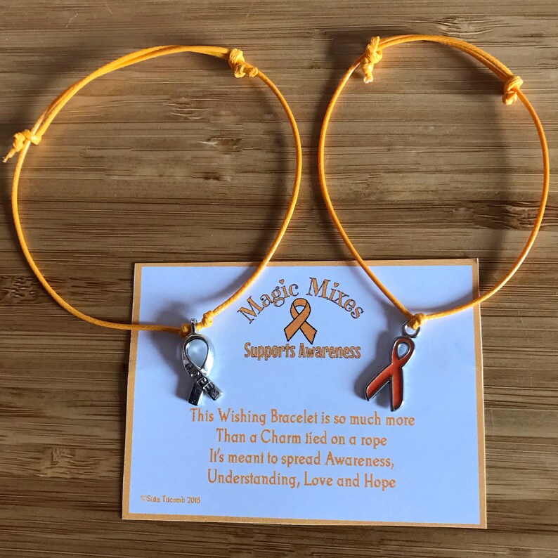 May include: Two orange cord bracelets with ribbon charms, one silver and one orange, rest on a wooden surface. A card reads "Magic Mixes Supports Awareness" and includes text about spreading awareness, understanding, love, and hope.